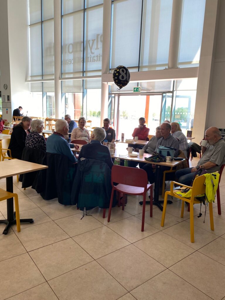 A group of carers chatting over coffee at café tables in the Plymouth Life Centre, a birthday balloon floating above them.