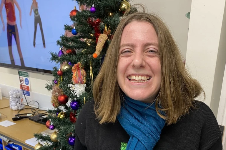 A woman with shoulder-length light brown hair, wearing a teal scarf and dark top, smiling broadly at the camera in front of a decorated Christmas tree, with a wall-mounted screen and desk visible in the background.