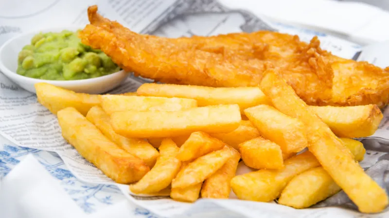 A fish and chip meal served on newspaper-print paper: battered fish, thick-cut chips and a small bowl of mushy peas.