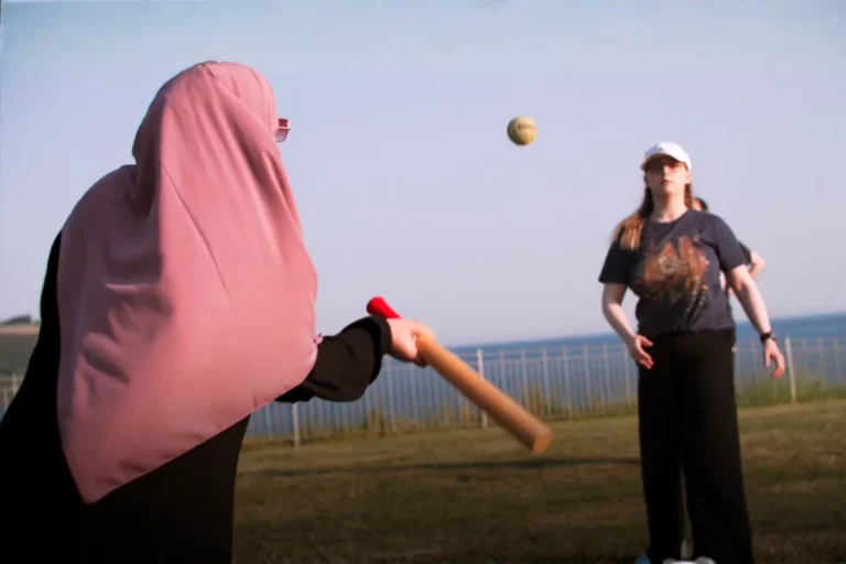 A person wearing a pink hijab and black clothing swings a baseball bat at an incoming ball while another person in a dark graphic t-shirt and white cap stands in the background on a grassy field near the coast, having just thrown the ball.