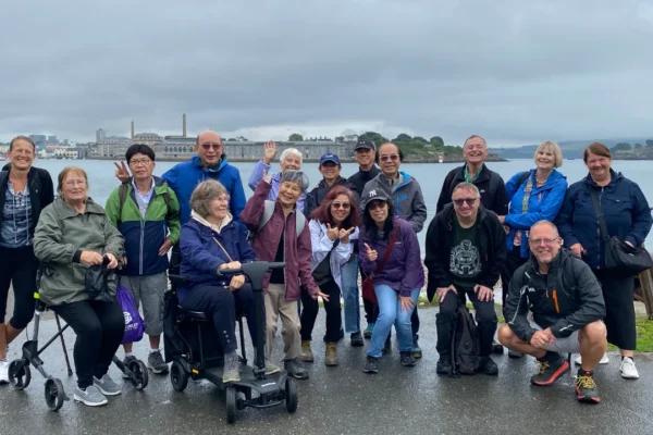 A group of approximately 18 people of various ages and ethnicities pose together for a photo on a wet waterfront promenade on an overcast day, with a harbor, historic buildings, and islands visible in the background. One person is seated in a mobility scooter, and the group is dressed in rain jackets and casual outdoor clothing, smiling and making playful gestures. The setting appears to be Plymouth, England, with what looks like the Royal William Yard and Drake's Island in the distance.