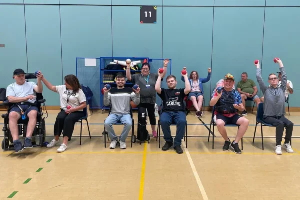 A group of approximately ten people sitting in chairs and a wheelchair inside a sports hall, smiling and raising bocce balls in the air. They are lined up against a light blue wall marked with the number 11, with sports equipment stored on a blue shelving unit behind them. The setting appears to be an inclusive bocce or adaptive sports session at a leisure center.