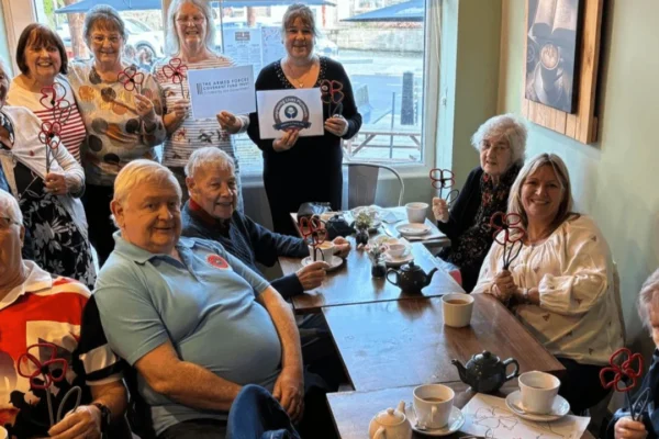 A group of approximately ten people, mostly older adults, gathered in a cozy café around wooden tables with teapots and cups, smiling at the camera while holding handmade red wire poppy crafts. Two women in the back are holding signs, one reading "The Armed Forces Covenant Fund Trust" and another displaying a circular logo. The setting appears to be a community craft and social event supporting a military-related charity.