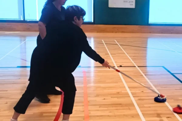 Two people in black clothing play floor curling in an indoor gymnasium, with one person lunging forward to push a curling stone using a long delivery stick while the other stands close behind offering support. The polished wooden floor is marked with colored lines, and a whiteboard and blue-tinted windows are visible in the background.