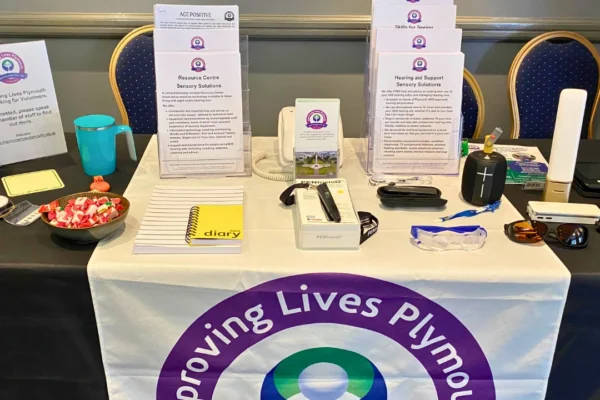 A display table for Improving Lives Plymouth, a charity supporting people since 1907, featuring informational leaflets about their Resource Centre Sensory Solutions and Hearing Aid Support services, along with assistive technology items including a PENfriend2 labeling device, magnifying glasses, sunglasses, a large-print diary, a portable speaker, and a bowl of sweets. A banner with the organization's purple and blue logo drapes over the front of the table, and a poster on the left advertises volunteer opportunities.