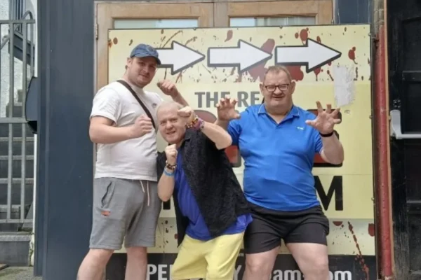 Three men posing playfully in front of a large sign with arrows reading "THE RE..." (partially obscured), appearing to be outside a horror or escape room attraction. The man on the left wears a white t-shirt, blue cap, and grey shorts while making fists; the man in the center is shorter, bald, and smiling while leaning forward; the man on the right wears a blue polo shirt and black shorts while making claw-like gestures with his hands. The sign behind them features blood splatter graphics and directional arrows. They are attending and escape room.