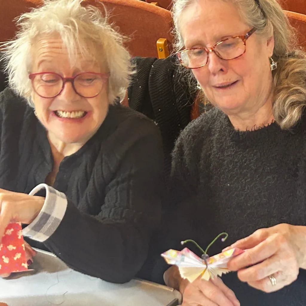 Two elderly women sitting together and smiling while doing crafts. The woman on the left, with white hair and red glasses, holds pieces of patterned fabric, while the woman on the right, with gray hair and tortoiseshell glasses, holds a handmade fabric butterfly with green pipe-cleaner antennae. Both are wearing dark clothing and appear to be enjoying a social crafting activity.