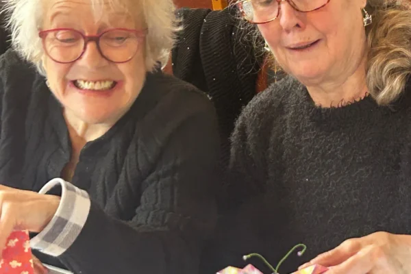 Two elderly women sitting together and smiling while doing crafts. The woman on the left, with white hair and red glasses, holds pieces of patterned fabric, while the woman on the right, with gray hair and tortoiseshell glasses, holds a handmade fabric butterfly with green pipe-cleaner antennae. Both are wearing dark clothing and appear to be enjoying a social crafting activity.