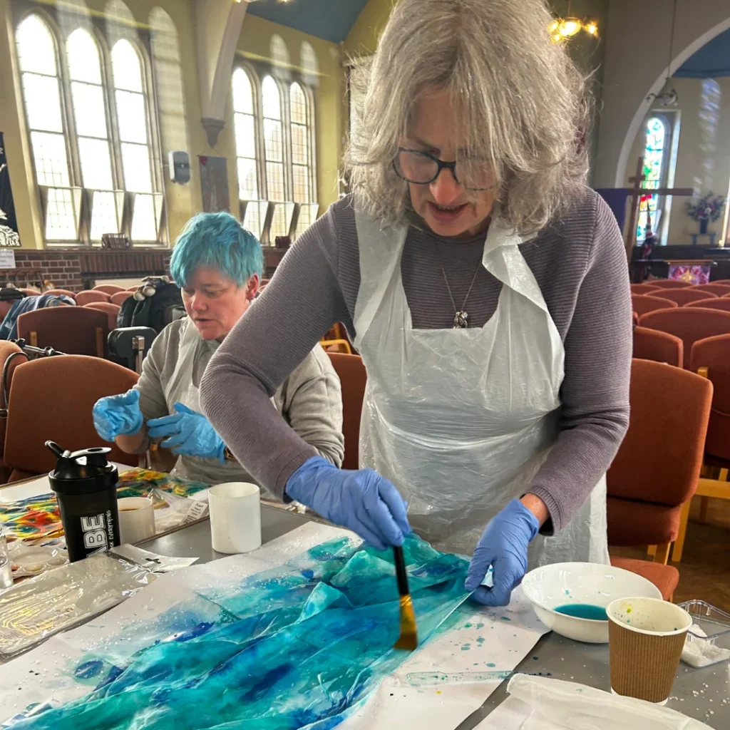 Two people creating art at a table inside a church hall. In the foreground, a woman with gray hair, glasses, and a plastic apron over a purple sweater uses a brush to apply turquoise and blue ink or paint onto a glossy surface, wearing blue nitrile gloves. Behind her, a person with short blue hair works on their own colorful piece, surrounded by cups, bowls of paint, and art supplies.