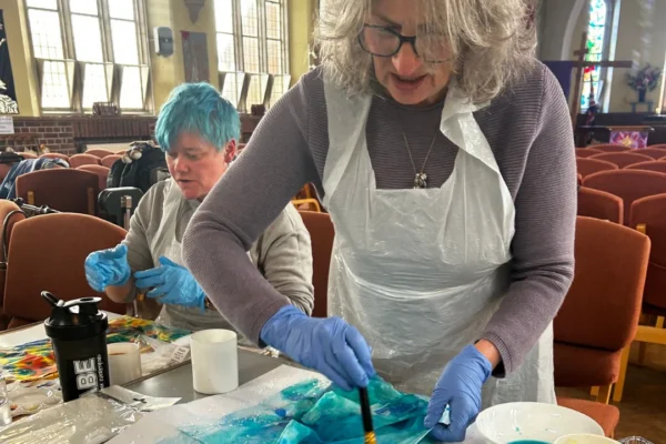 Two people creating art at a table inside a church hall. In the foreground, a woman with gray hair, glasses, and a plastic apron over a purple sweater uses a brush to apply turquoise and blue ink or paint onto a glossy surface, wearing blue nitrile gloves. Behind her, a person with short blue hair works on their own colorful piece, surrounded by cups, bowls of paint, and art supplies.