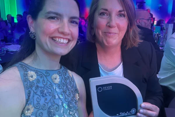 Two women posing together at a formal awards ceremony with colorful green and blue lighting. The woman on the left wears an embellished light blue dress and smiles broadly, while the woman on the right in a dark blazer holds a Devon Chamber award trophy for Collaboration of the Year 2025, awarded to Plymouth Wellbeing Hub Network.