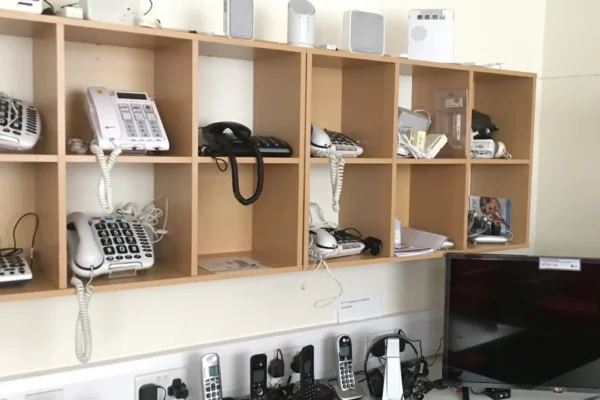 A display wall of assistive telephones and communication devices, featuring multiple corded and cordless phones with large buttons designed for people with hearing or vision impairments, arranged on wooden cubby shelves. Below the shelves, additional cordless handsets sit on a counter alongside a flat-screen TV, remote controls, and various electronic accessories. It is a resource center for assistive technology.