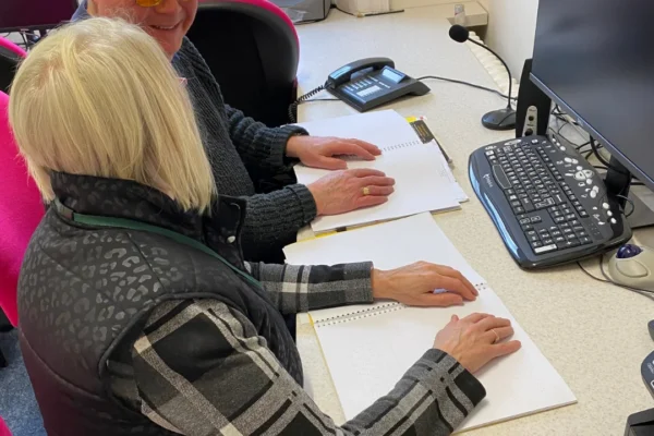 Two people sit at a desk reading Braille documents together. A bald man wearing yellow-tinted glasses smiles while running his hands across a Braille page, while a woman with blonde hair reads from a separate spiral-bound Braille book beside him. The desk is equipped with a computer monitor, keyboard, mouse, and desk phone in what appears to be an office or resource center.