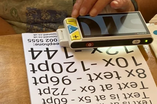A person's hand holds a Sight A Clover 5 portable electronic magnifier over a printed text size demonstration sheet showing various magnification levels from 0.8x to 12x. The handheld device has a small screen, yellow plus and minus zoom buttons, and a green power indicator light. The sheet beneath displays sample text at increasing sizes from standard 12pt up to 144pt, with the device positioned over the larger text samples.