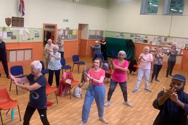 A group of mostly older women participate in a group exercise or tai chi class in a community hall, following the movements of an instructor wearing a dark outfit and cap in the lower right. The hall features a wooden floor, blue and orange chairs pushed to the sides, a Union Jack flag on the wall, and notice boards along the green and orange painted walls.