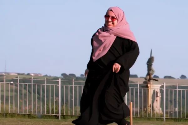 A smiling woman wearing a pink hijab, sunglasses, and a flowing black abaya walks across a grassy area. Behind her is a white metal fence with rolling green hills and trees in the background under a clear blue sky. A small statue or ornamental figure is visible near the fence to the right.