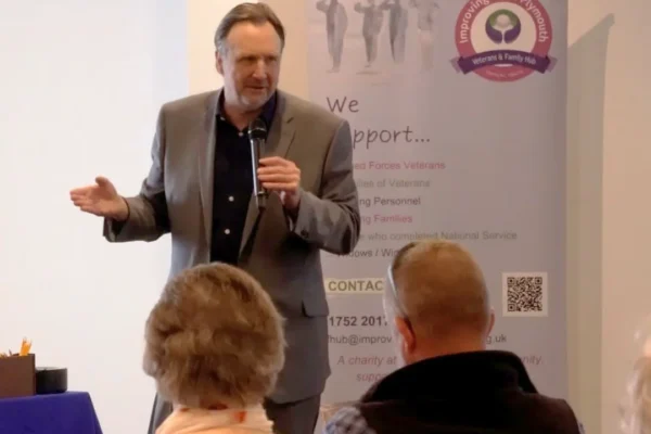 A man in a gray blazer and dark shirt speaks into a microphone while addressing a small seated audience at an Improving Lives Plymouth Veterans & Family Hub event. The pull-up banner behind him lists support services for armed forces veterans, their families, serving personnel, and widows. Several audience members, including an older woman in a floral top and a man in a plaid shirt, are visible from behind.