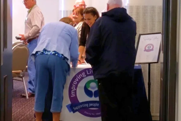 Several people gathered around a registration table at a charity event for an organization whose banner reads "Improving Lives, Supporting People" with a charity number partially visible. A young woman behind the table assists attendees, while a seating plan is displayed on an easel to the right. The scene is viewed through a doorway of what appears to be a conference or banquet venue.