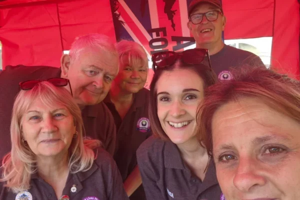 A group of six smiling people take a selfie under a red pop-up tent, wearing matching dark gray polo shirts with Improving Lives Plymouth Veterans and Family Hub logos. The woman on the left has a name badge reading Ellie, and a Union Jack banner with soldier silhouettes hangs behind them, suggesting a veterans charity event or outreach stall.