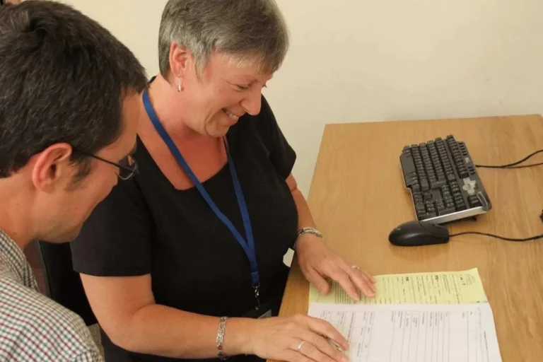 Woman with short gray hair wearing a blue lanyard smiles while reviewing paperwork with a man in glasses at a desk with a keyboard and mouse, appearing to be in a consultation or advisory meeting.