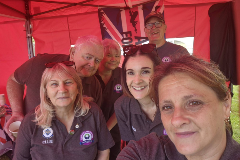 Group selfie of six smiling people from Improving Lives Plymouth Veterans & Family Hub wearing matching dark gray polo shirts with organizational badges, standing under a red pop-up tent with a Union Jack military banner behind them. One name tag reads "Ellie."