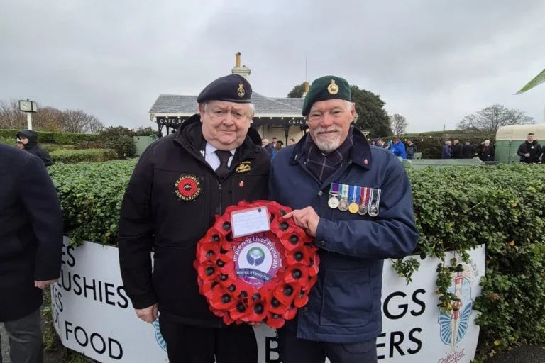 Two military veterans holding a red poppy wreath from Improving Lives Plymouth Veterans & Family Hub at a Remembrance Day ceremony, one wearing a "We Remember Submariners" badge with navy beret and the other wearing a Royal Marines green beret with service medals.