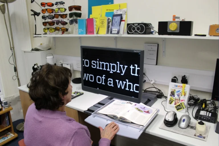 Woman using a magnifier to read a magazine, with enlarged white text displayed on a black screen, surrounded by low vision aids including tinted glasses, large print diaries, and assistive devices on shelves.
