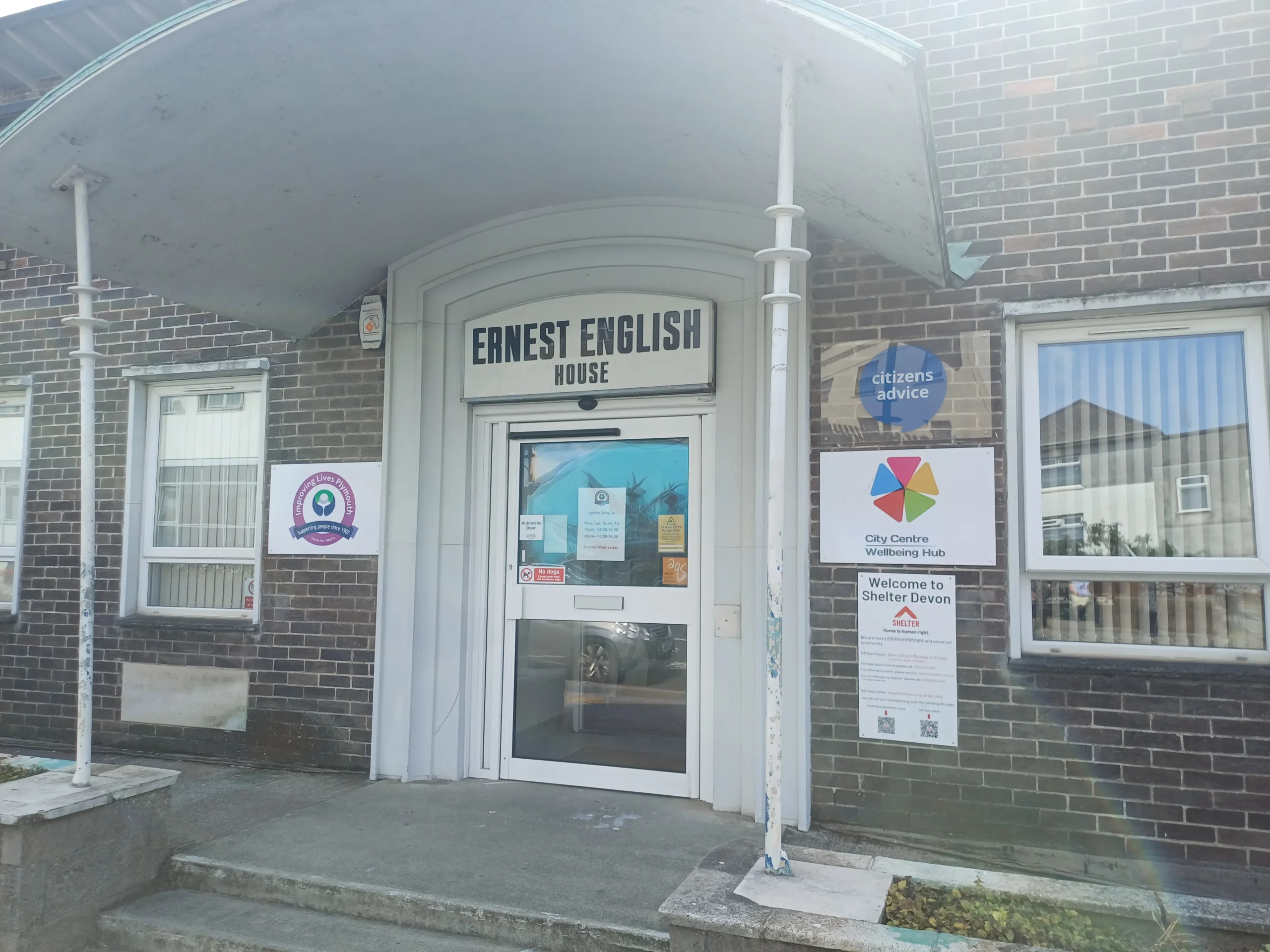 The entrance to Ernest English House in Plymouth, a brick building with a curved white canopy over automatic doors. Signs beside the entrance show the Improving Lives Plymouth logo, Citizens Advice, the City Centre Wellbeing Hub and Shelter Devon.