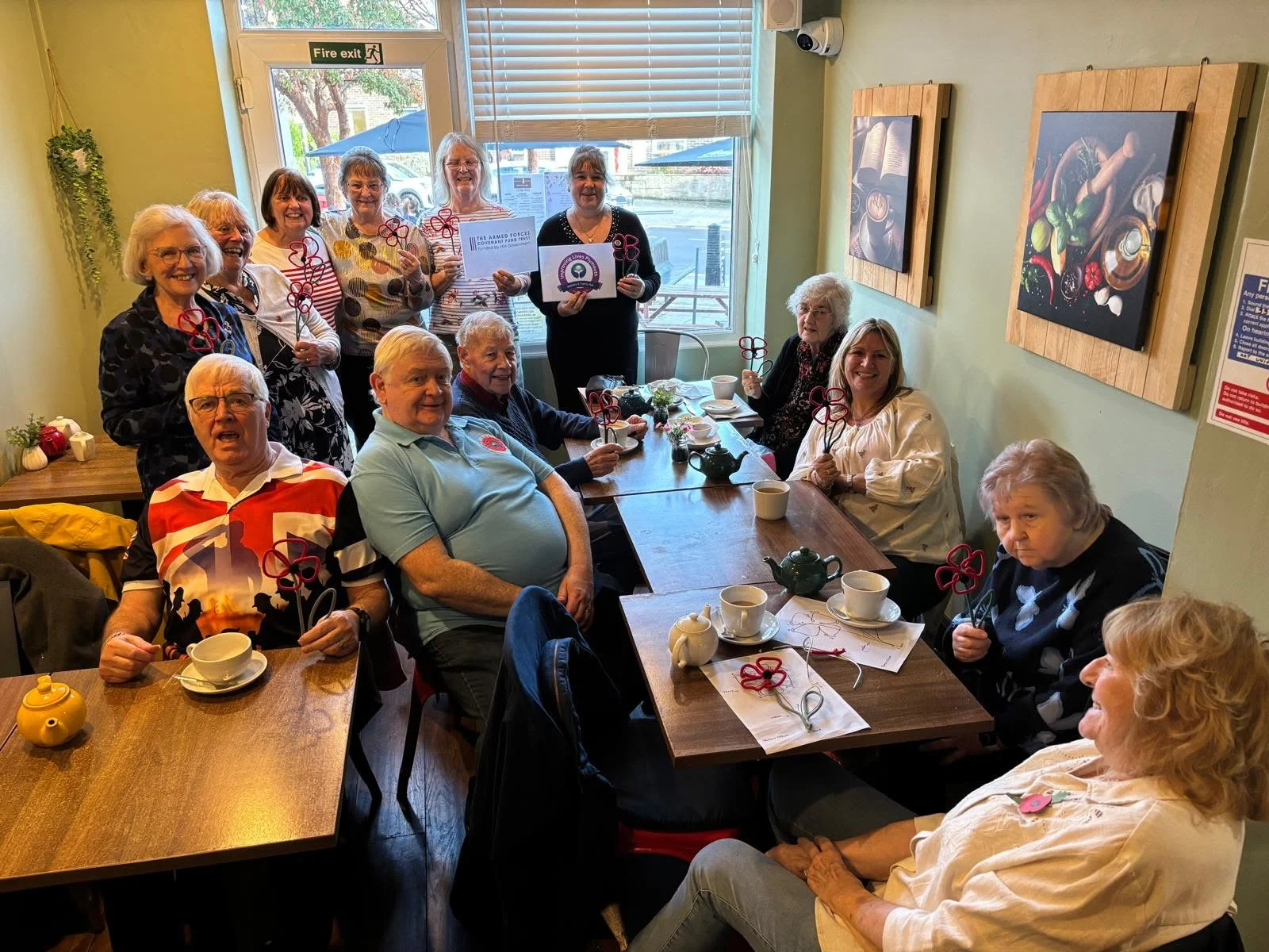 A group of around fourteen people smile in a small cafe, holding handmade poppies crafted from red and grey pipe cleaners. Two women at the back hold up signs for the Armed Forces Covenant Fund Trust and Improving Lives Plymouth, and teapots and cups fill the tables.