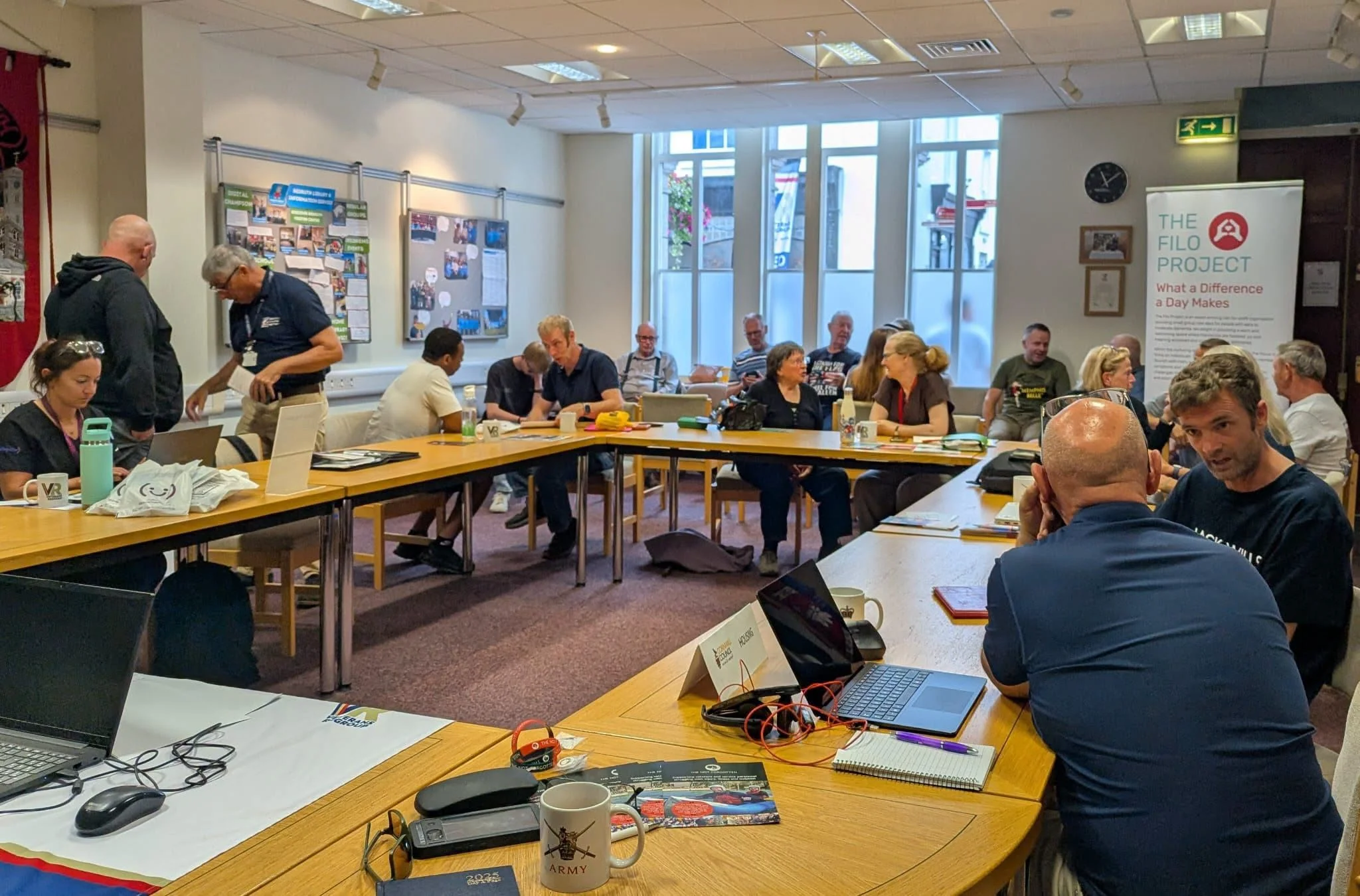 Around twenty people chat in small groups at tables arranged in a large rectangle in a bright meeting room at Redruth Library. Veterans Regroup mugs and leaflets sit on the tables, alongside an Army mug and a pop-up banner for The Filo Project.