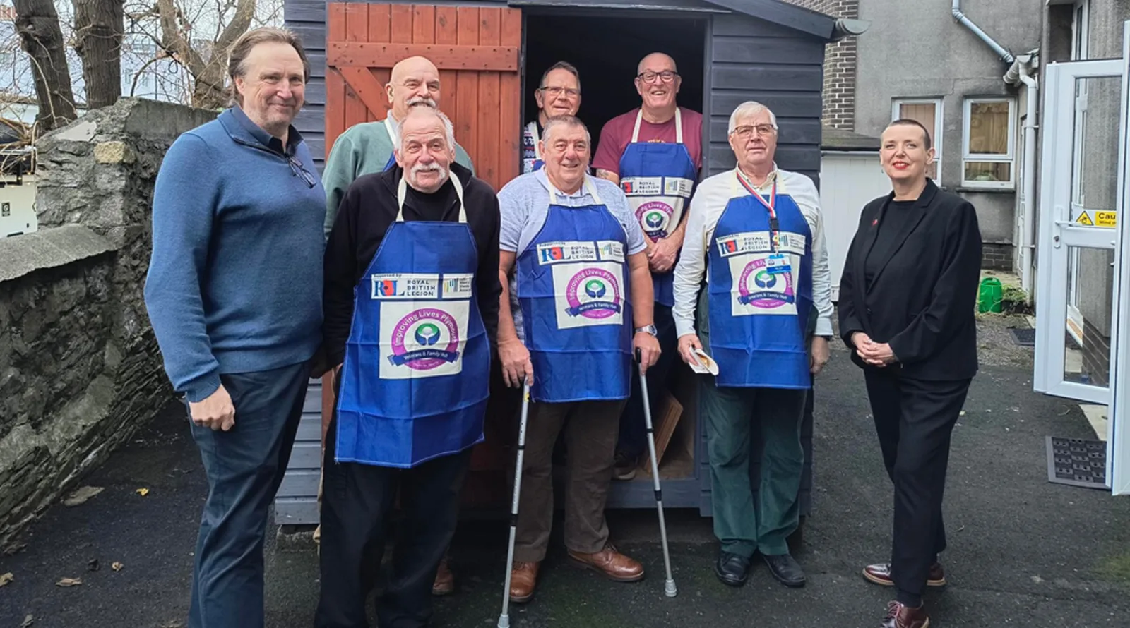 Eight people smiling outside a wooden shed, six of them in blue Improving Lives Plymouth Veterans and Family Hub aprons, one leaning on crutches, with two others alongside them.