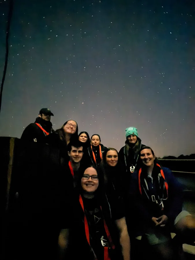 Nine young people and staff in life jackets crowd together on deck at night under a sky full of stars, smiling at the camera.