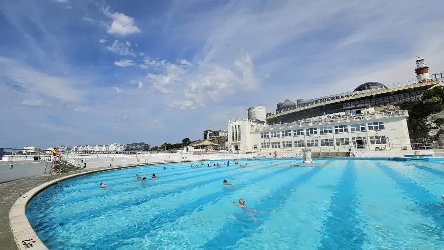 A wide view of Tinside Lido on a sunny day, swimmers dotted across the pale blue pool, the white art deco lido building and Smeaton's Tower lighthouse on the cliff behind.