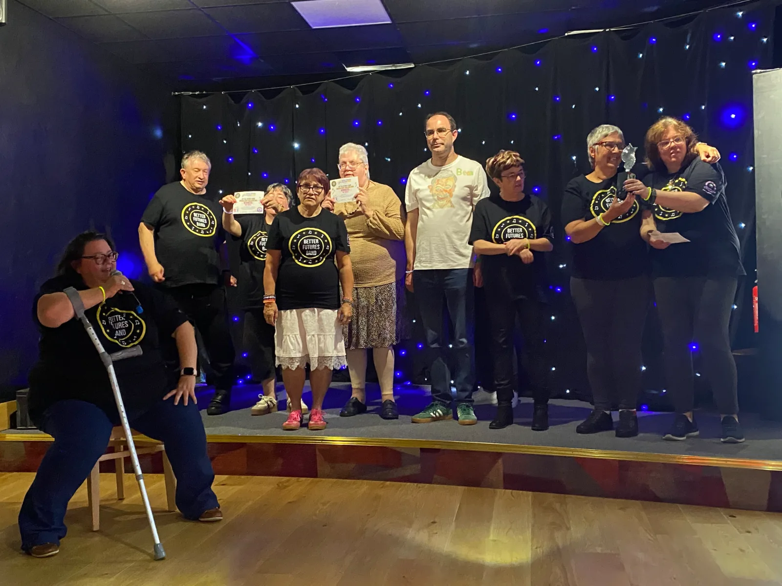 The Better Futures Band lined up on the small stage at the Cattedown Social Club in front of a black star-cloth curtain, most in black band t-shirts, two holding up certificates and one holding a trophy, while a woman at the front sings into a microphone.