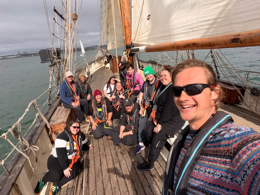 Twelve young people and staff in orange-strapped life jackets smile up at the camera from the wooden deck of the sailing ship Tectona under full sail, with Plymouth Sound and the city shoreline behind them.