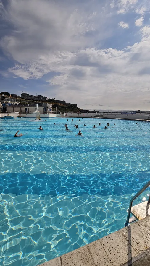Swimmers spread across the far end of Tinside Lido near the fountain, with the Hoe and Royal Citadel walls behind under a bright, cloudy sky.