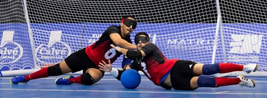 Two goalball players in eyeshades dive across a blue indoor court to block a ball in front of the goal.
