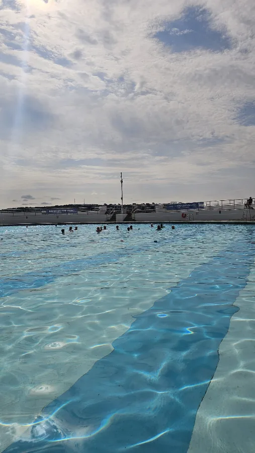 A group of swimmers gathered together in the water at the far side of Tinside Lido, backlit by a bright overcast sky, sunlight rippling across the pool.