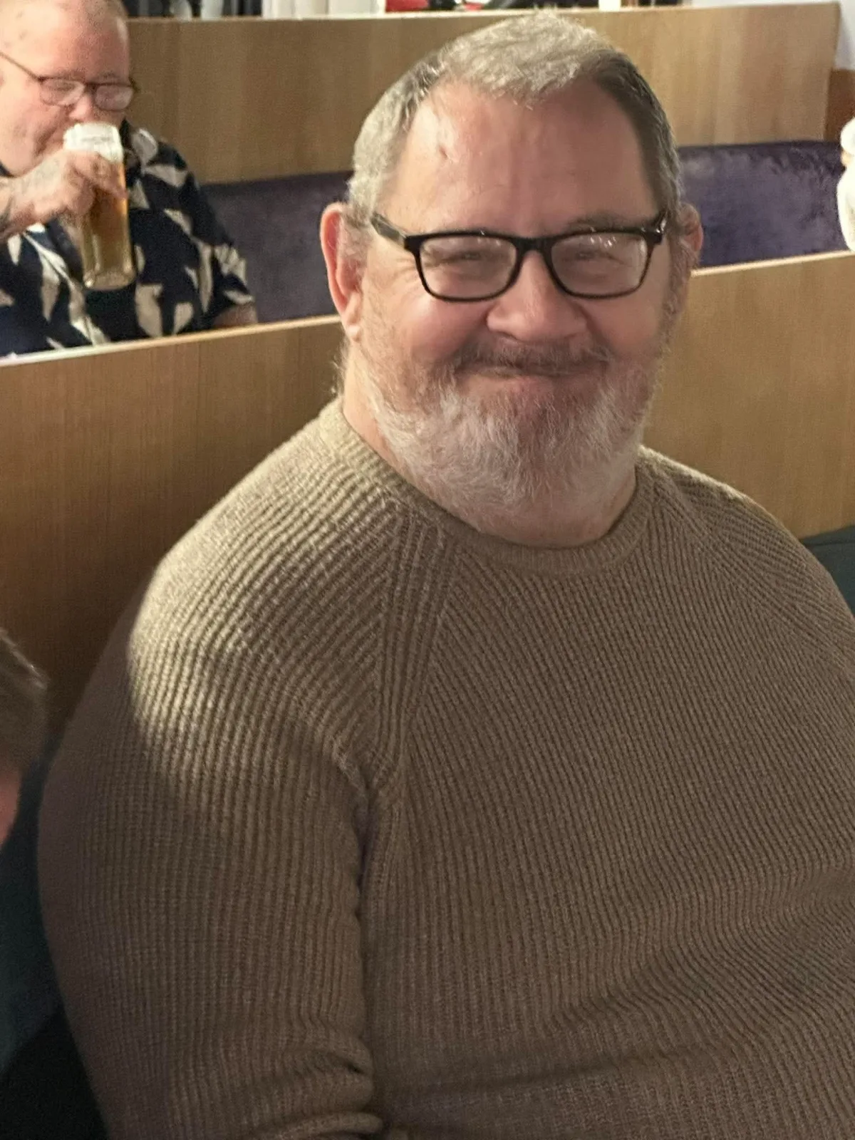 A man with a grey beard and glasses smiles warmly at the camera in a ribbed brown jumper.