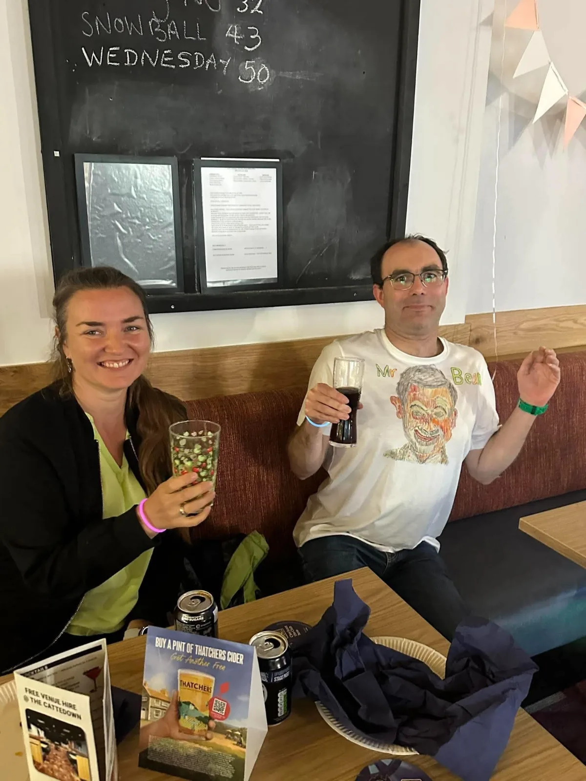 Two guests sitting on a bench seat raising their drinks to the camera, one wearing a hand-drawn Mr Bean t-shirt.