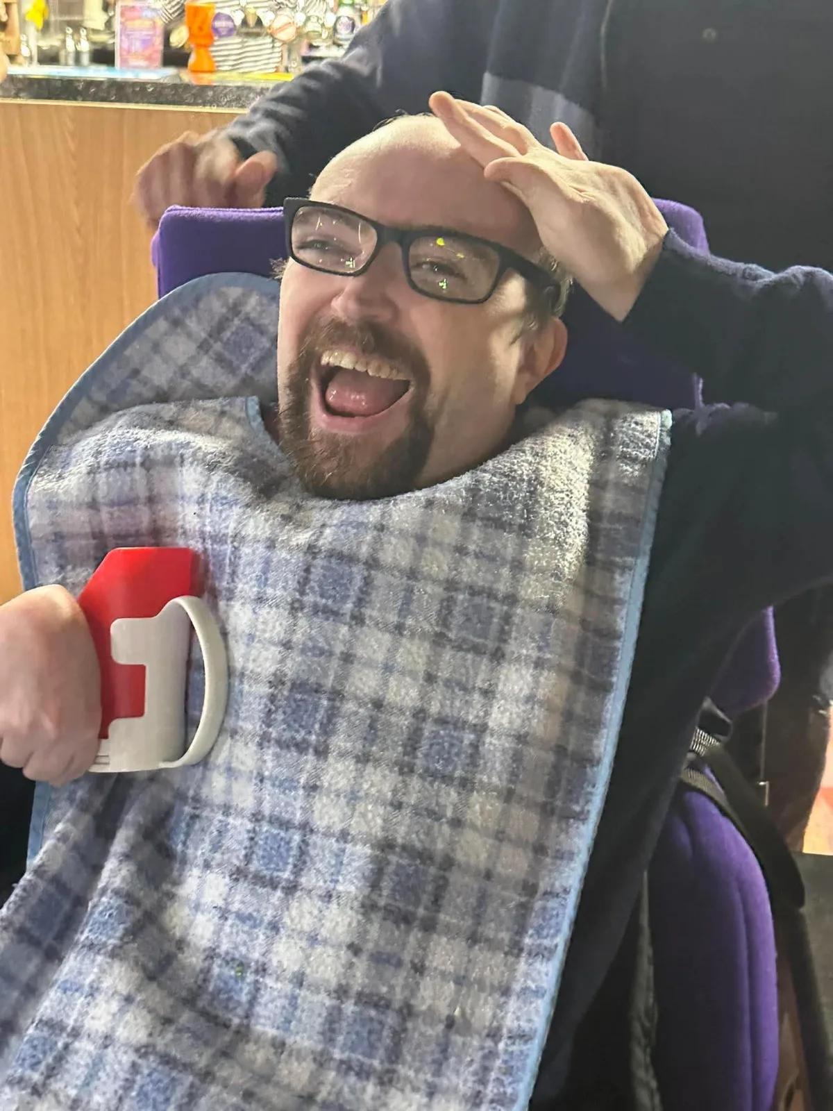 A guest in glasses laughs with delight, one hand on his head and a red cup in the other.