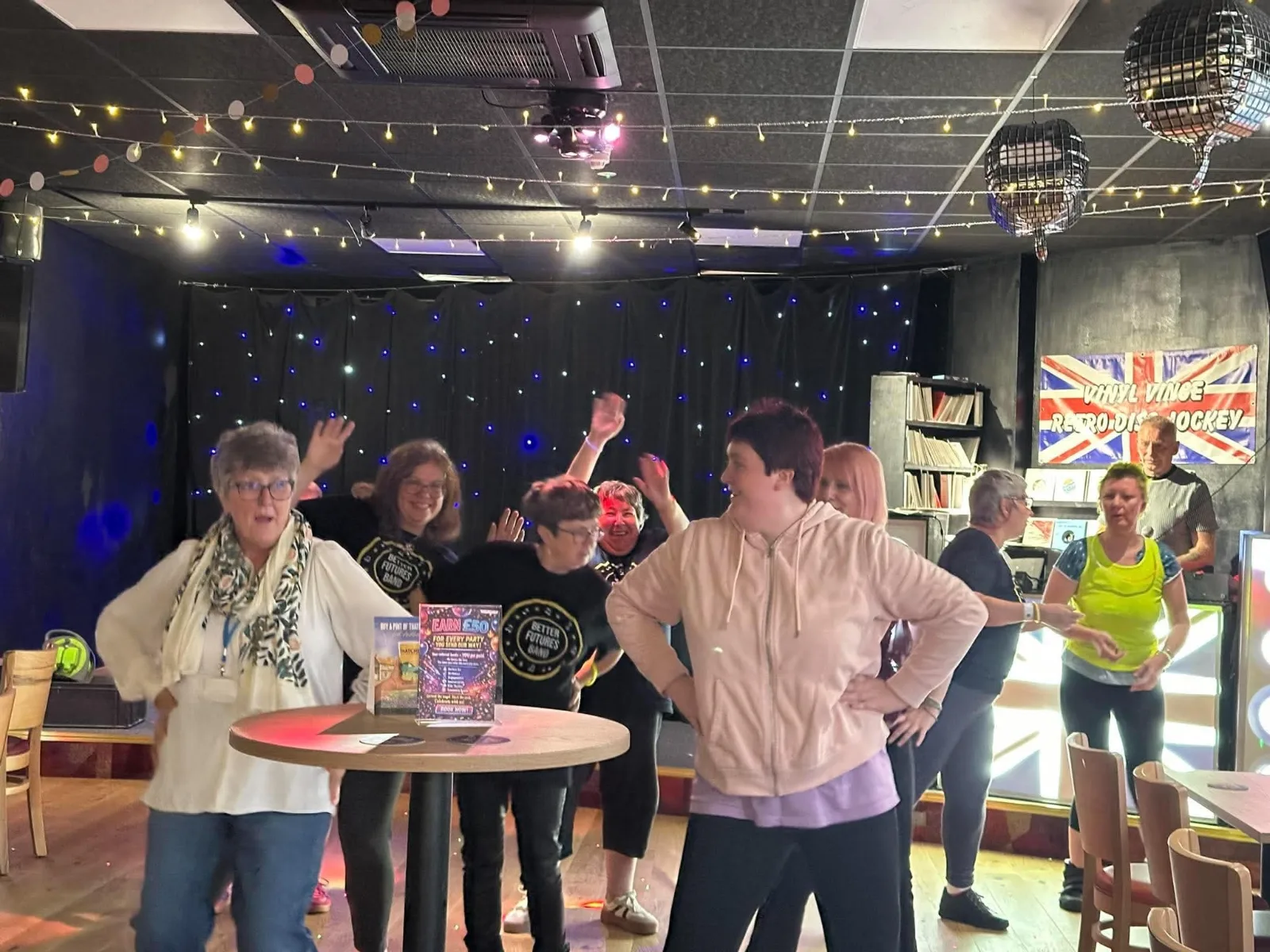 Guests dancing with arms raised under fairy lights and disco balls at the Cattedown Social Club, a Vinyl Vince Retro Disc Jockey banner on the wall behind.