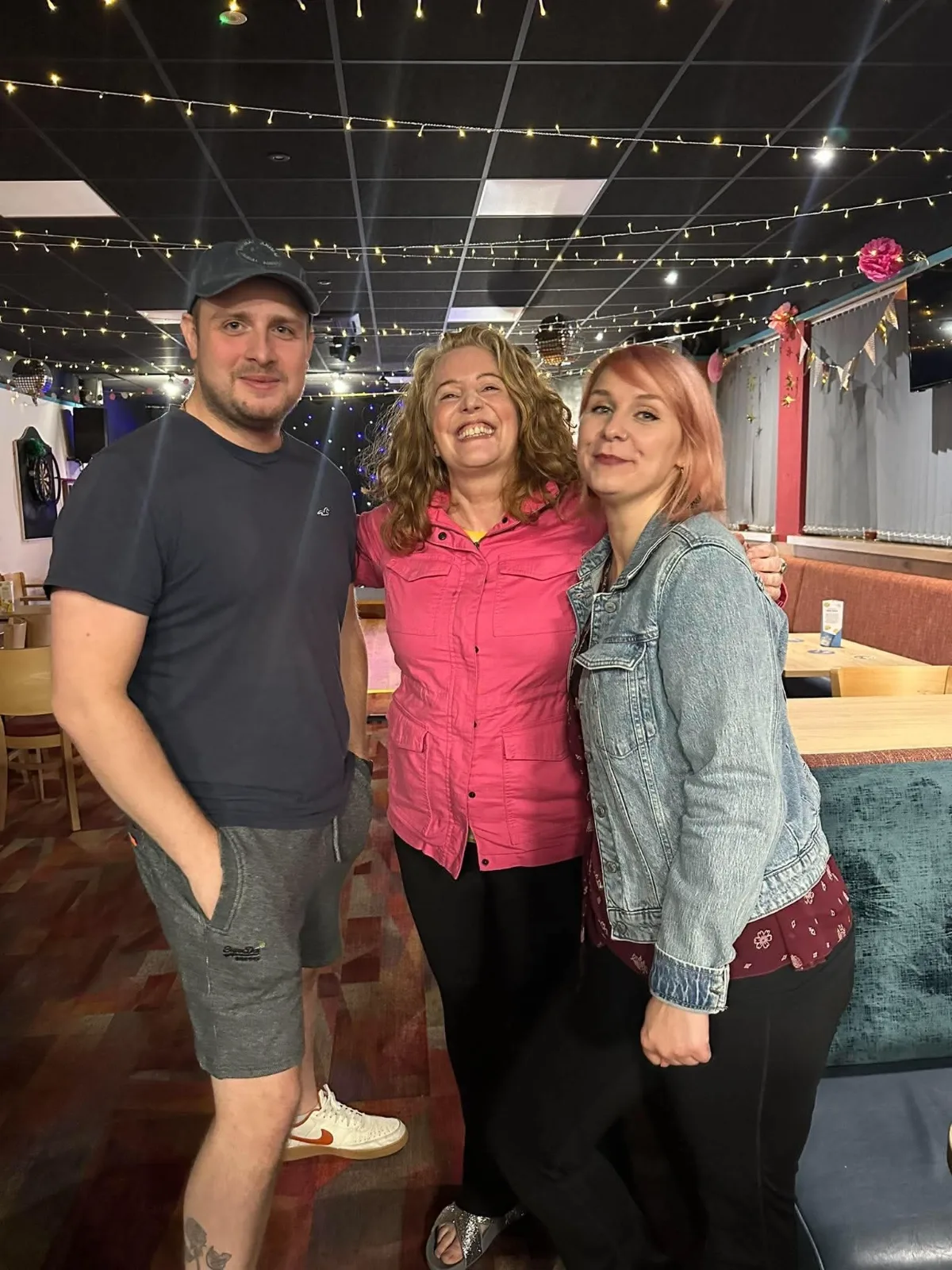 Three friends stand arm in arm under fairy lights, the woman in the middle laughing.
