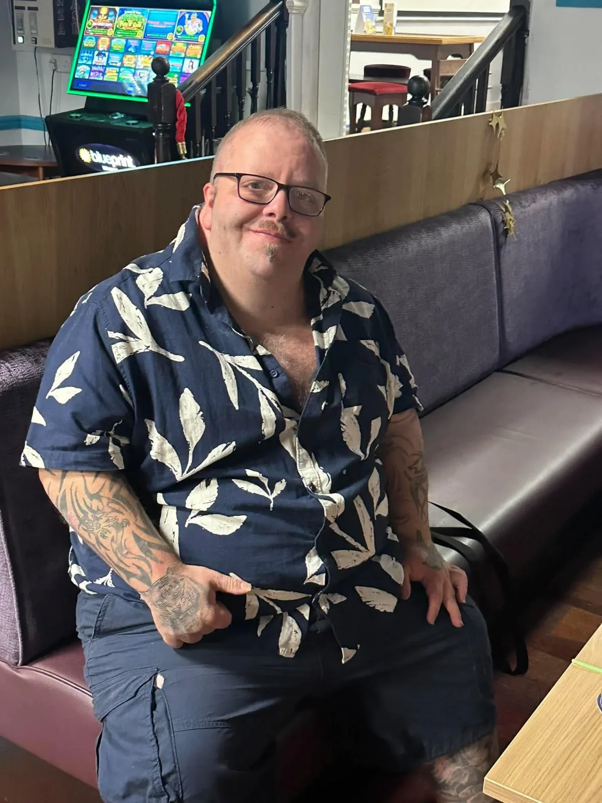 A man in a navy leaf-print shirt sits on a bench seat and smiles at the camera.