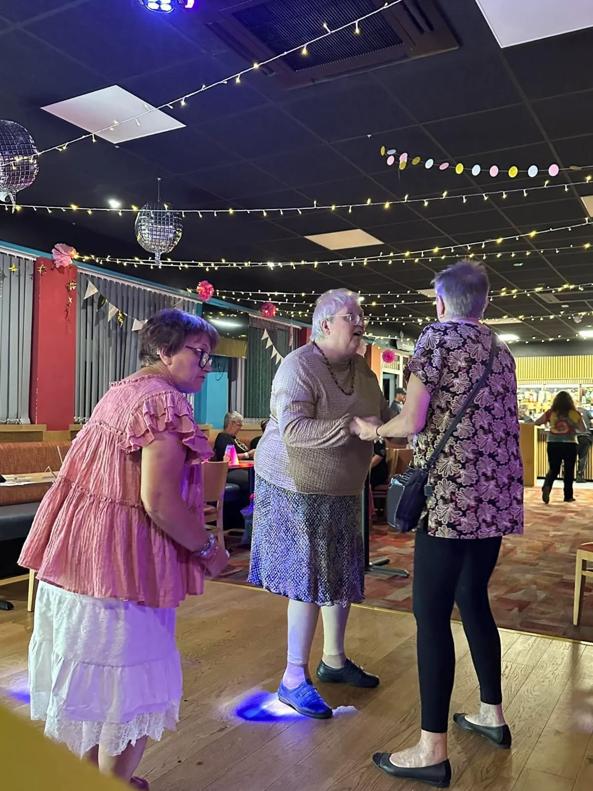 Three women dancing together on the dance floor under strings of fairy lights and disco balls.