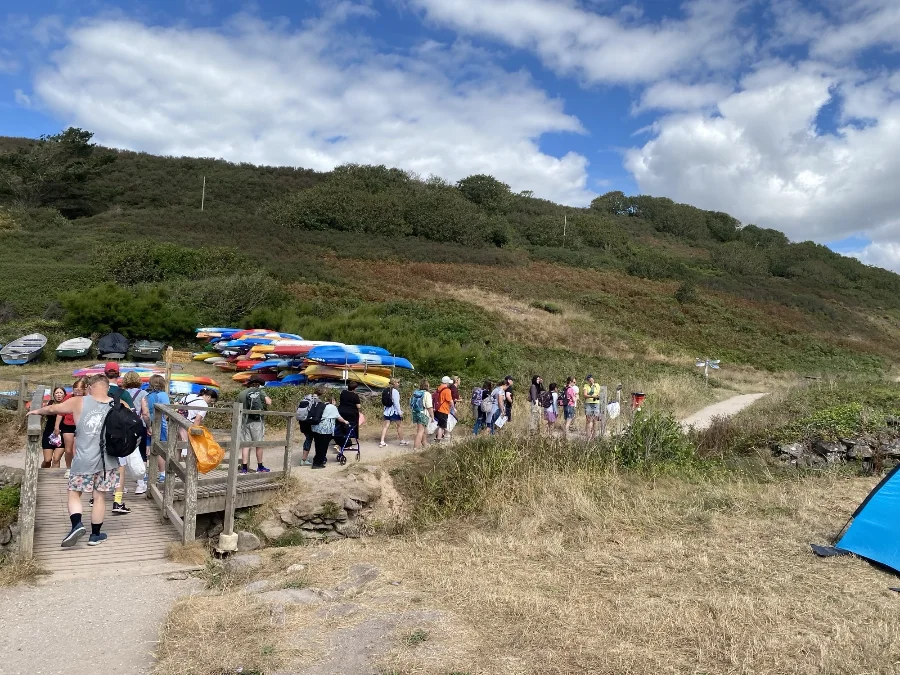 The group walks in a line along a coastal path past a wooden footbridge and stacked kayaks, under a blue sky with white clouds.