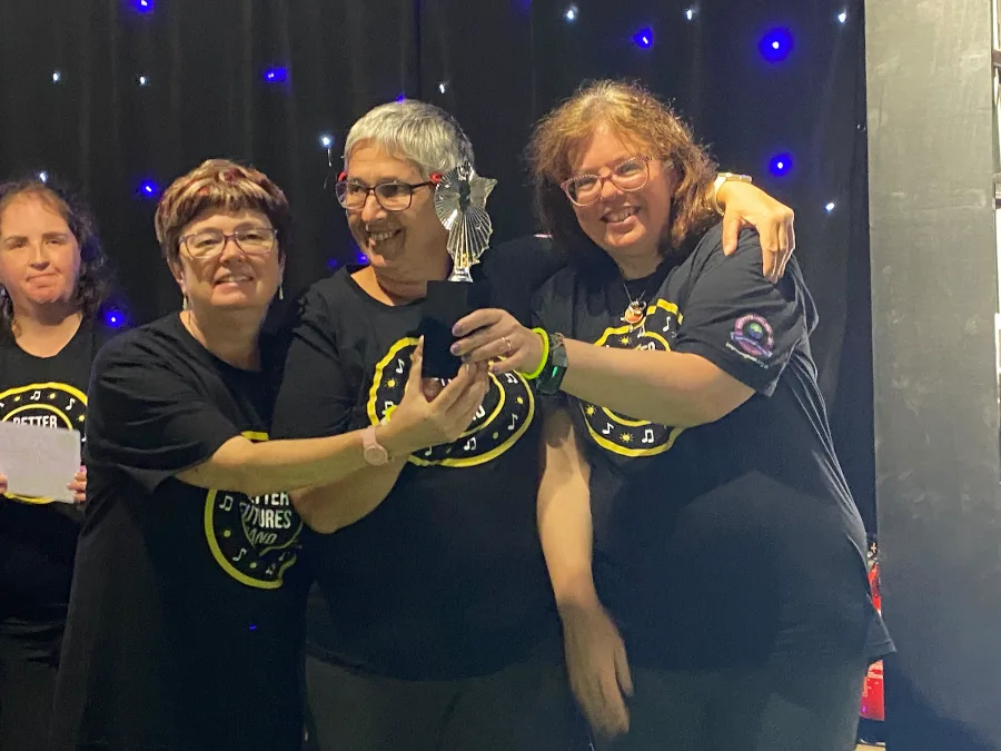 Three smiling women in Better Futures Band t-shirts hold a star-topped trophy together in front of a star-cloth curtain.